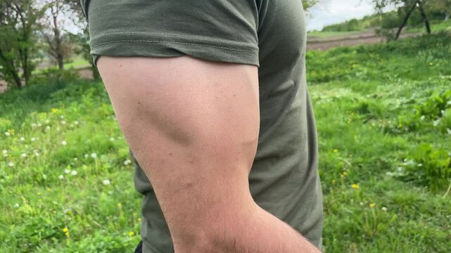 Young man in a green t shirt flexes biceps repeatedly in a grassy field, showcasing strength and fitness in nature
