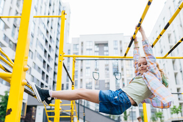 Kid, adventure and fun on jungle gym park, balance or energy for physical growth. Cute school girl climbing and hanging on outdoor horizontal bar with modern building on background.