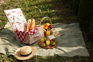 Picnic basket with tableware, different snacks, juice and hat outdoors, space for text