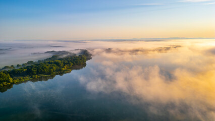 A tranquil morning unfolds over Rozmberk Pond, where soft mist blankets the water's surface, reflecting the warm hues of dawn. The surrounding greenery adds to the serene beauty of this location.