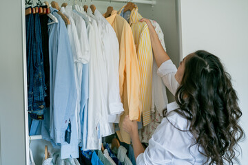 Young dark-haired curly beautiful woman is sorting things in large spacious closet. Home organization. Washing things. Analysis of clothes by season.