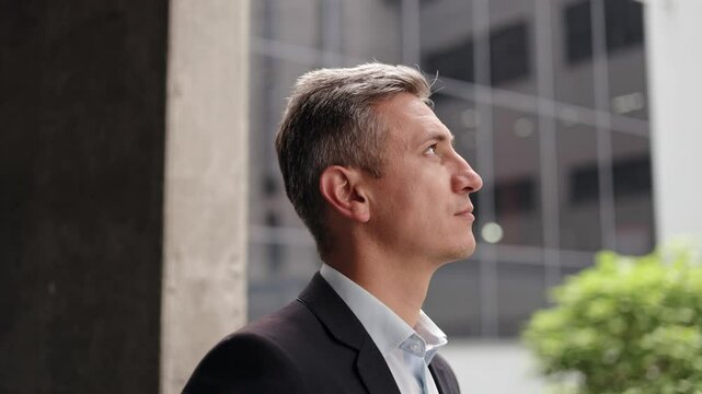 A middle-aged businessman with grey hair, wearing a suit, stands in a modern city center, looking thoughtfully into the distance. He projects an image of business success, leadership, ambition.