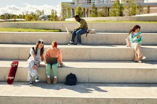 Wide angle shot of students of diverse ethnicity sitting on concrete seats of amphitheater in park using laptops and phones surfing net after classes outdoors