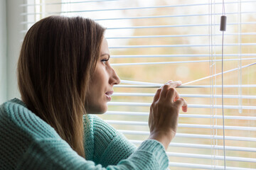 Young Woman peering through blinds sitting indoors on a sunny day, lost in thought and reflection...