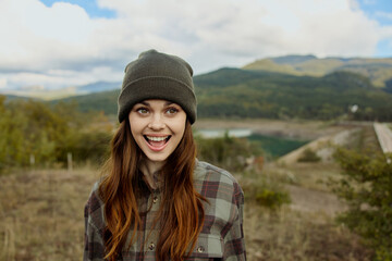 Serene young woman in plaid shirt and beanie standing by lake with mountain background contemplating the beauty of nature