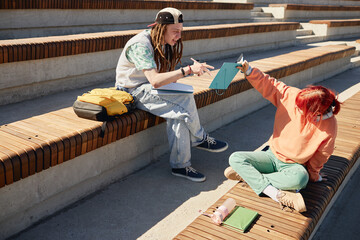 Laid back female college student sitting on bench showing teenage boy notebook with drawings and male friend lively commenting on it, casual outdoor learning scene, copy space