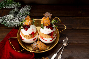Two glasses of Christmas dessert with gingerbread cookies, whipped cream and cranberries on dark wooden background