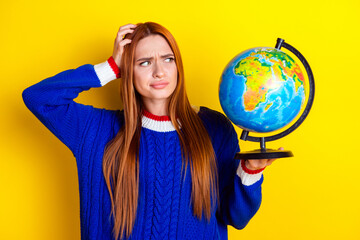 Photo portrait of pretty young girl look confused hold globe wear trendy blue knitwear outfit isolated on yellow color background