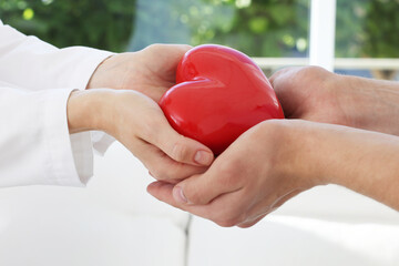 Doctor giving red heart to patient at white table in clinic, closeup