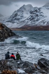 Three People Sit On Rocks Watching The Ocean Waves Crash Against The Shore In Front Of A Snow Covered Mountain Range