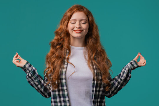 Tranquil woman relaxing, meditating, refuses stress. Ginger lady breathes deeply, calms down, blue studio background. Yoga, moral balance, zen concept.