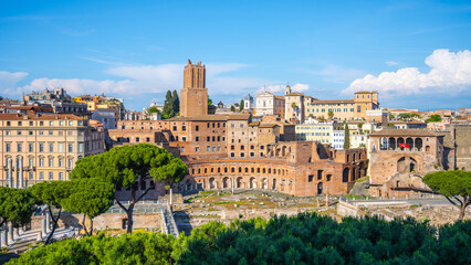 Naklejka premium The ancient Trajan Market stands proudly in Rome, Italy, showcasing its remarkable architecture while surrounded by blooming greenery and majestic ancient ruins under a bright blue sky.