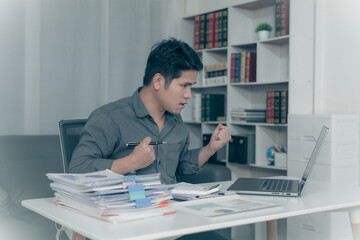 A young Asian businessman working from home is delighted with the good news, raises his hands and is excited about the success of his business.