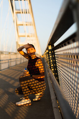 Stylish woman enjoys a sunny afternoon on a bridge, sipping water while basking in the golden glow...