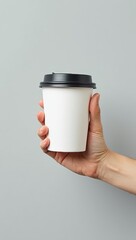 Close-up of a hand holding a white disposable coffee cup with a black lid against a gray background.