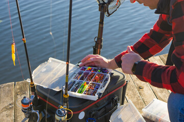 A fisherman selecting a lure from an organized tackle box on a wooden pier during a sunny autumn...