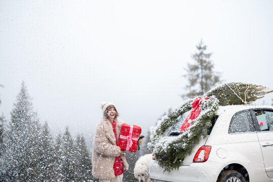 Portrait of happy woman stands with a gift box on snow storm while traveling by car decorated with Christmas wreath and tree. Concept of happy winter holidays and magic