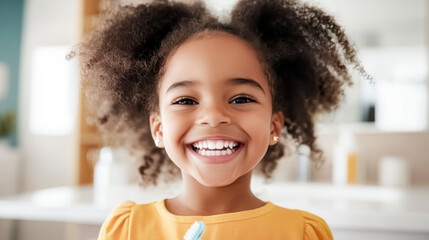 A young girl with curly hair is happily brushing her teeth in a bathroom with a white tile wall and a potted plant in the background.