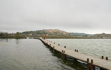 Wooden walkway coming out from the right of the image surrounded by water with a town on the horizon and the sky with grey clouds
