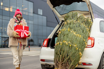 Young cheerful woman in winter clothes stands with a gift box near shopping mall and Christmas tree in front. Concept of shopping for winter holidays