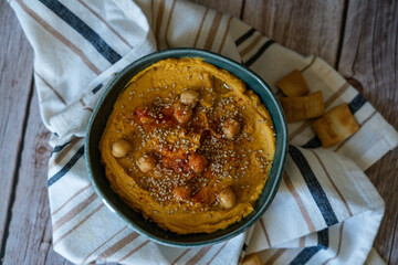 A creamy and vibrant pumpkin hummus garnished with chickpeas, presented on a dark background that accentuates the warm, rich colors of the dish. The rustic presentation is enhanced by a simple cloth, 