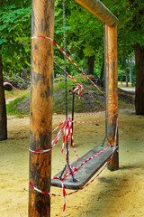 Fototapeta premium Wooden swing on chains on empty playground in park. White and red warning tape. Prohibited to use