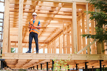 Fototapeta premium Carpenter constructing wooden frame two-story house near the forest. Man in glasses holding hammer, dressed in protective helmet and orange safety vest. Concept of ecological modern construction.