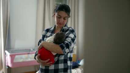 Mother gazing lovingly at her newborn, nestled in her arms in a warm, homey bedroom. The baby is...