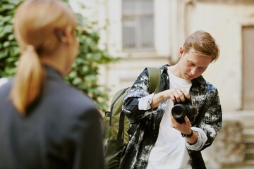 Young man holding camera and making adjustments while standing in outdoor setting with greenery in background and another person nearby focusing on him