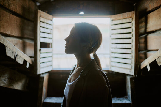 Silhouette of a woman standing in front of open wooden shutters in a rustic setting giving a sense of calm and introspection
