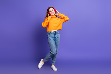 Photo of blonde cheerful teenager girl in orange turtleneck and jeans hang out dancing listen headphones isolated on purple color background