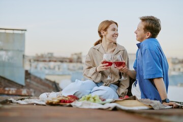 Couple smiling while holding wine glasses and enjoying picnic on rooftop with cityscape backdrop at sunset. Fruits and cheese placed on blanket while sharing laughter