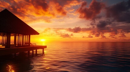 A serene water villa stands on stilts over the turquoise waters of the Indian Ocean. The sky explodes in vibrant hues of orange, pink, and purple as the sun dips below the horizon, casting a golden gl