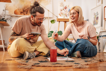 Artistic couple sitting on atelier floor and painting together on canvas
