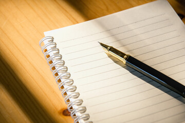 Close-up of a notebook and pen on a wooden table with sun shining