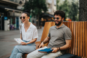 Two happy colleagues enjoying a conversation while seated on an outdoor bench, holding notebooks and smiling.