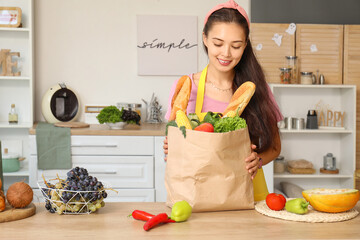 Beautiful young Asian woman with paper bag full of fresh products at table in kitchen