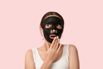 Surprised young woman with activated charcoal mask on her face against pink background