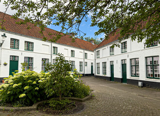 Naklejka premium Brugge, Flanders, Belgium - June 22, 2024: White facades and red roofs of historic residential houses around courtyard of Wevershof with flowering plants and tree foliage