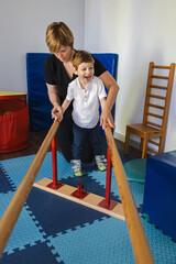 Young male child is engaging in physical therapy, assisted by a caregiver, using parallel bars to...