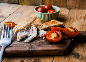 Plate with rustic food, fork, and knife on a wooden board. The plate contains chicken, bread, and tomatoes.