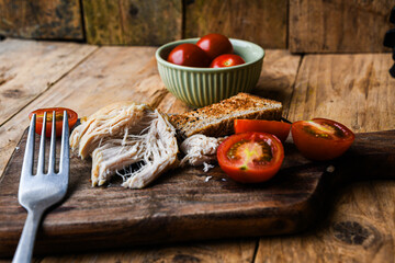 Plate with rustic food, fork, and knife on a wooden board. The plate contains chicken, bread, and tomatoes.