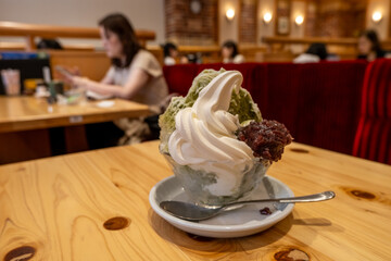 A bowl of soft-serve vanilla and matcha-flavored ice cream with red bean paste is placed on a table, ready to eat, with a background of several Asian people seated in a cozy and casual café.