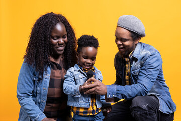 Proud parents listening to their infant son singing a song on microphone, laughing and being adorable together against yellow background. Happy african american family of three enjoy music.