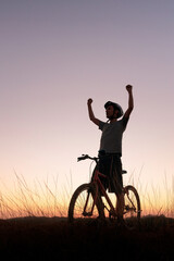 Mountain biker pauses at dusk, raising their arms in victory. The scenic sunset and tranquil...