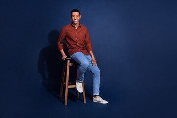 Full length photo of cheerful positive guy dressed brown shirt sitting stool emtpy space isolated...