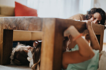 Two children giggle and hide under a sturdy wooden table while playing a joyful game of hide and...
