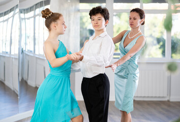 Motivated adolescent ballroom dancers, girl and boy in performance outfit practicing elegant dance moves in pair in bright studio, with female coach supervising in background