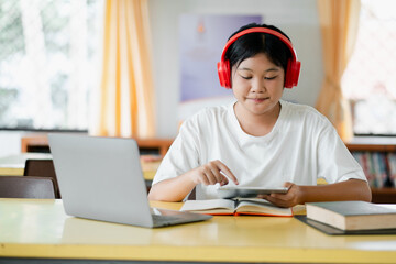 A young student wearing red headphones uses a tablet in a classroom setting, with a laptop and books on the desk, focused on online learning.