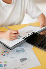 Close-up of a person reviewing financial documents with charts and graphs, using a pen and a laptop on a yellow desk.